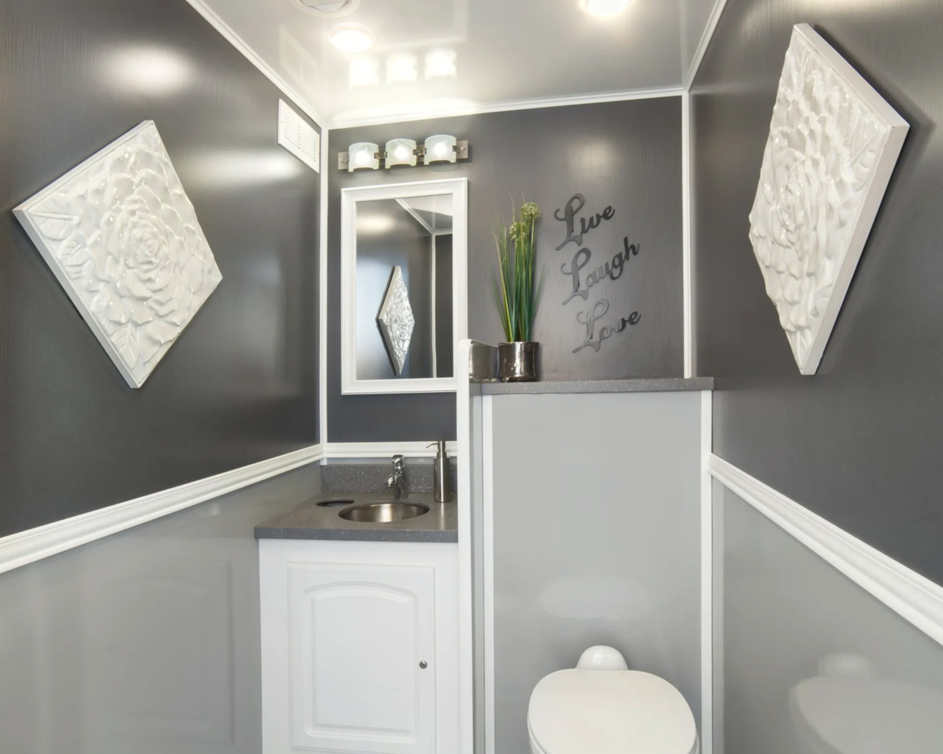 Restroom Trailer featuring gray walls, decorative white wall art, a mirror, a small sink with a faucet, and a white toilet. The decor includes the words Live Laugh Love and a small plant in a metallic pot.
