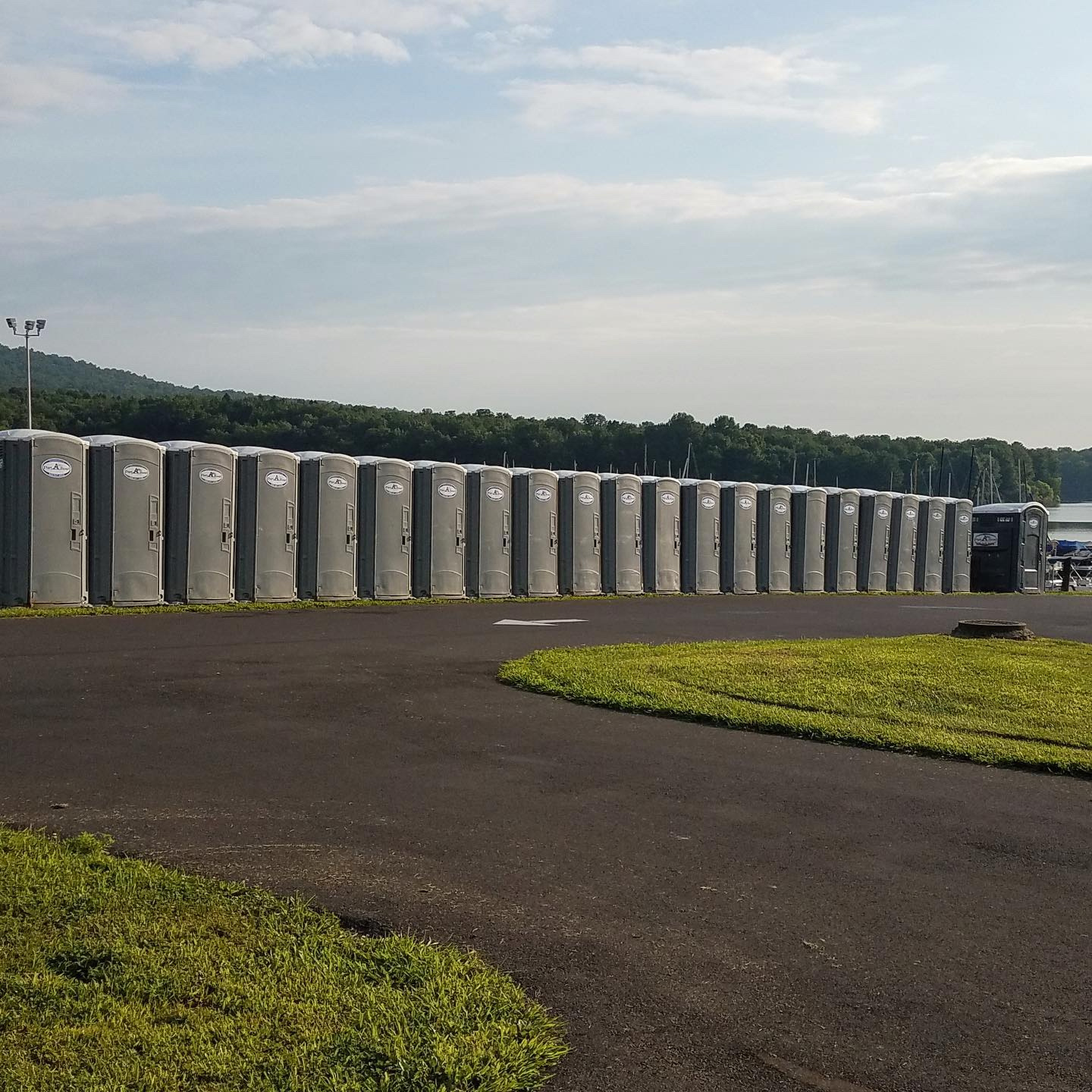 A row of portable toilets lined up along a paved area, with a grassy patch nearby and a view of trees and a waterbody in the background, under a partly cloudy sky.