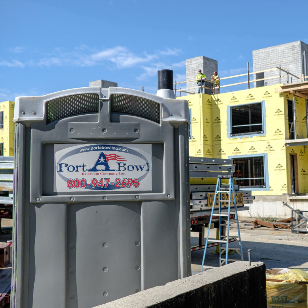 A gray portable toilet with the brand name Port Bowl is in the foreground, while a construction site with workers and partially built structures is visible in the background under a clear blue sky.