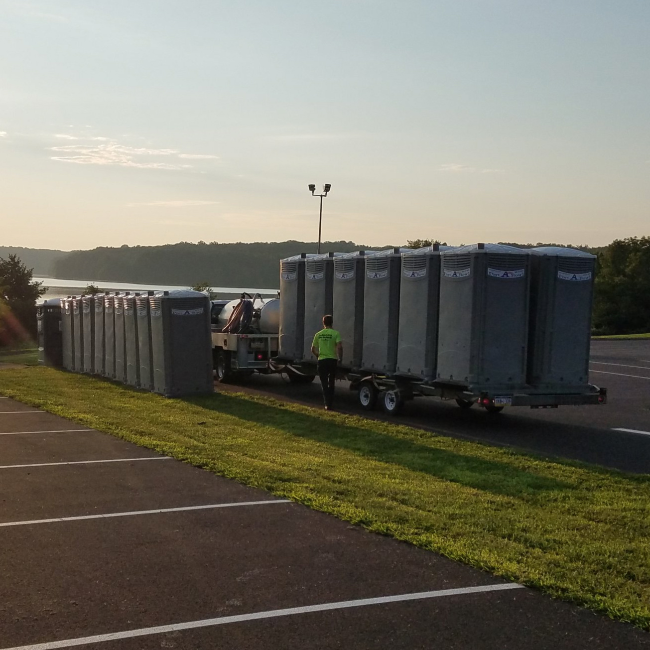 A line of portable toilets is arranged on a trailer, with a worker wearing a bright green shirt walking beside it. The setting is near a calm body of water, with lush greenery in the background and a soft morning light illuminating the scene.