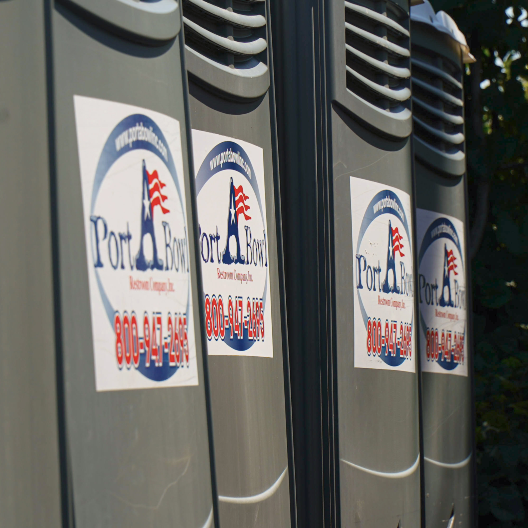 Rows of portable toilets with a company logo and contact information clearly visible. The toilets are gray and have ventilation slats at the top.