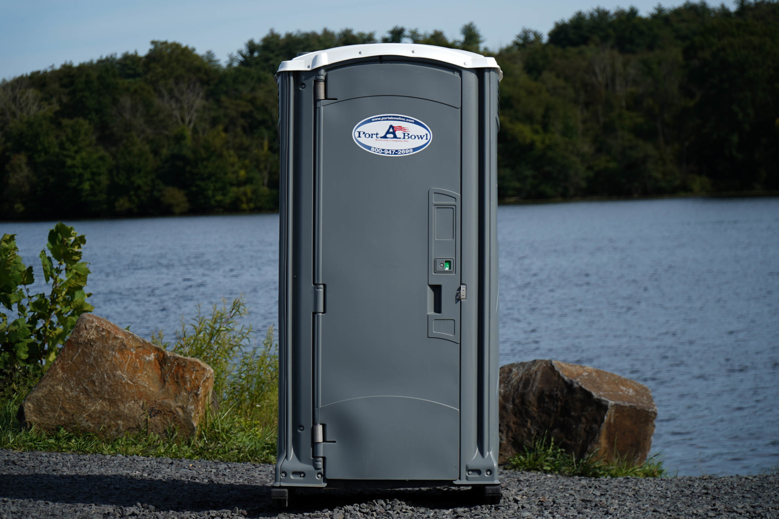 Portable Toilet located near a body of water, surrounded by greenery and rocks, with a clear sky in the background.