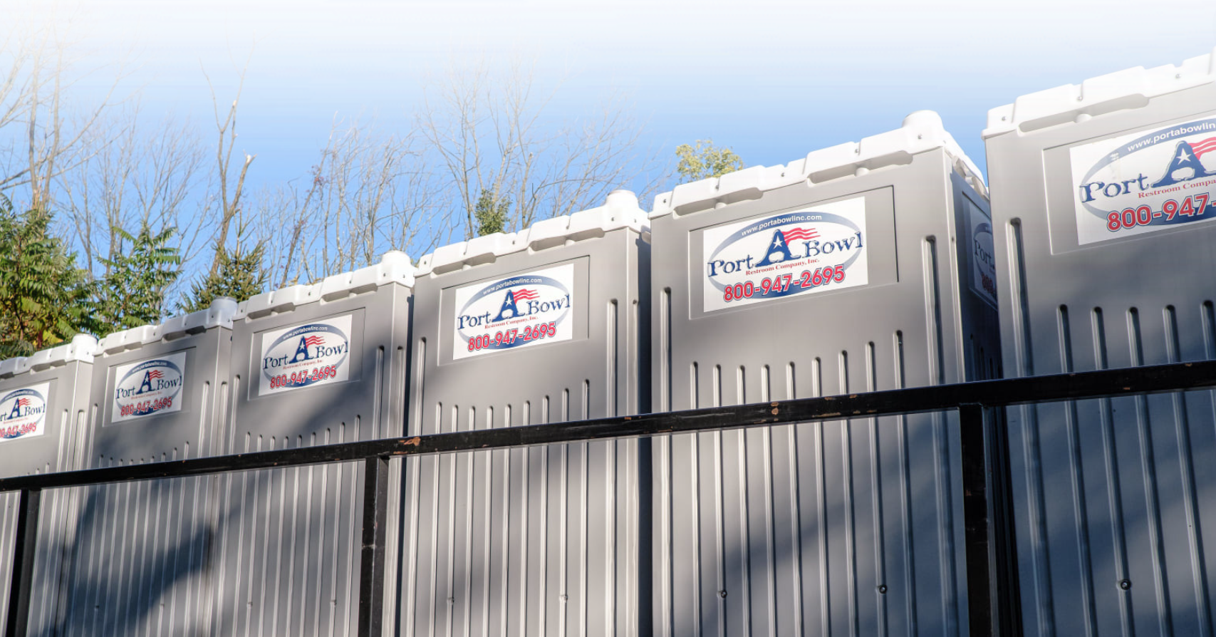 A row of portable toilets with a gray exterior, showcasing the logo and contact information for Port-A-Bowl Restroom Company. The units are lined up against a wooden fence, set in a wooded area with trees in the background.