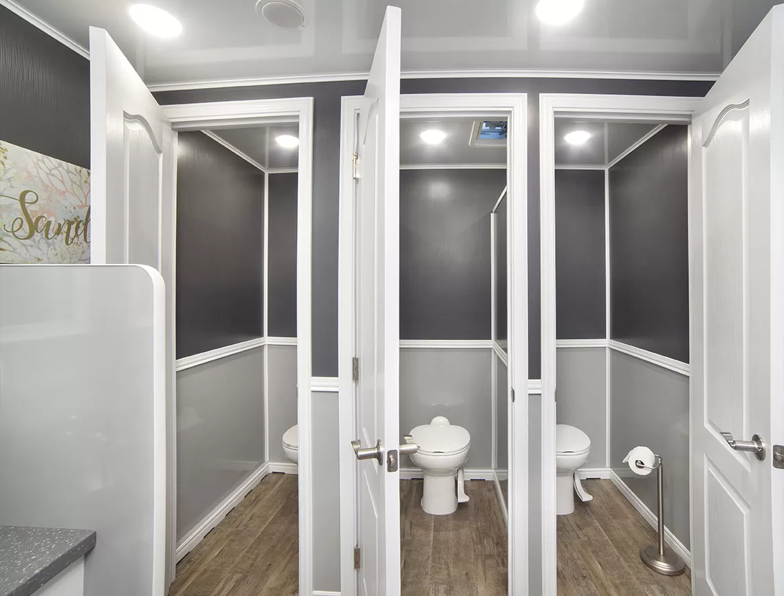 Restroom Trailer featuring multiple private stalls with white doors, two modern white toilets, and grey walls accented with white trim and flooring. The ceiling has recessed lighting, and there is a decorative wall sign.